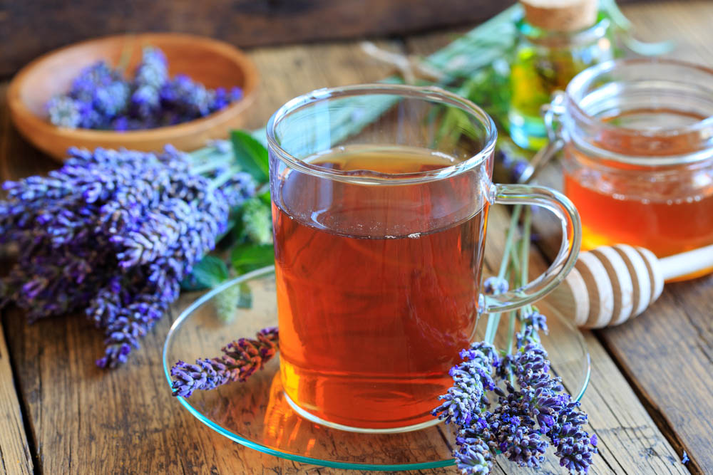 Taza de té de lavanda sobre un plato de vidrio, rodeada de flores de lavanda frescas y frascos de miel sobre una mesa rústica de madera