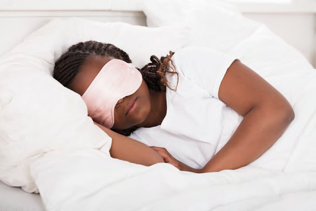 Niña durmiendo con un antifaz rosa sobre una cama blanca, abrazando la almohada mientras descansa plácidamente