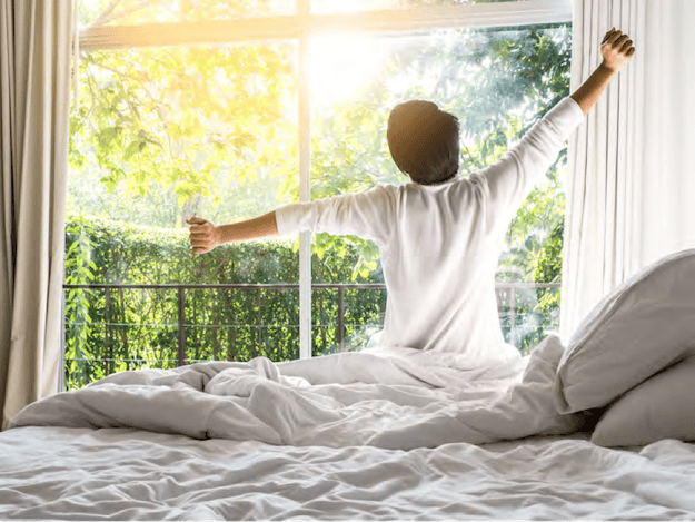 Persona sentada en la cama estirándose frente a una gran ventana con luz de la mañana y vistas a un jardín verde