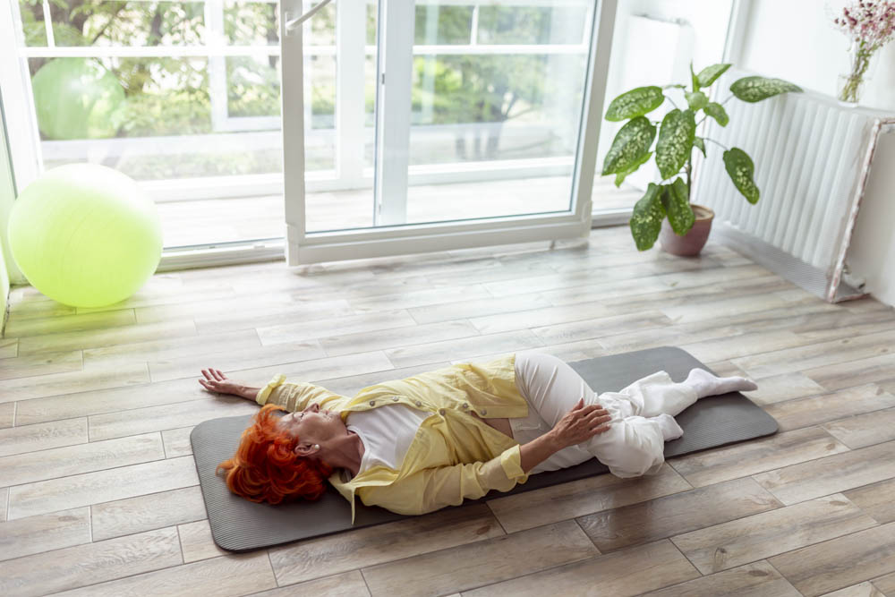 Mujer mayor de cabello rojo vibrante acostada de lado sobre una esterilla de yoga, vestida con un top amarillo y pantalones blancos, en una habitación luminosa con grandes ventanas y una pelota de ejercicio verde cerca