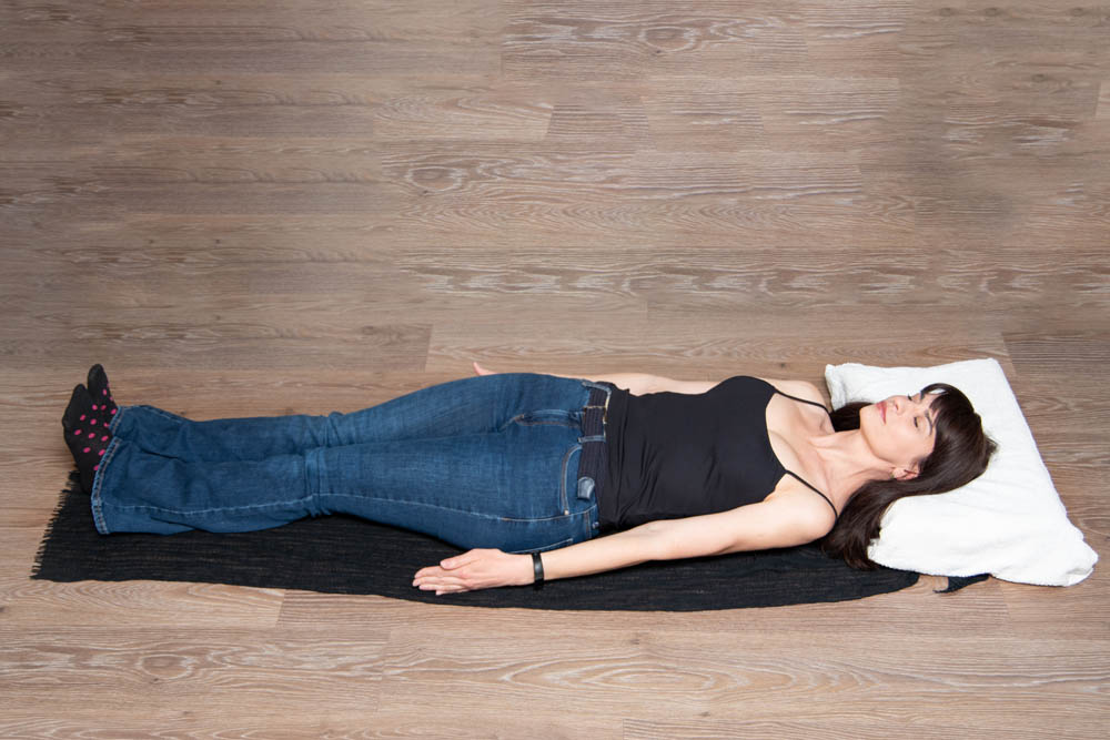 Mujer acostada boca arriba sobre una esterilla de yoga negra, vestida con camiseta sin mangas negra y jeans azules, con los brazos extendidos y una almohada bajo la cabeza, en una posición tranquila y relajada sobre un suelo de madera