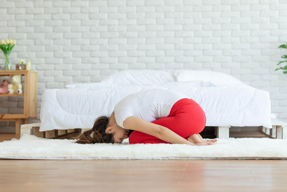 Mujer en una postura de yoga con la frente apoyada sobre las rodillas, sentada sobre una alfombra suave en una habitación luminosa con una cama al fondo, creando un ambiente tranquilo