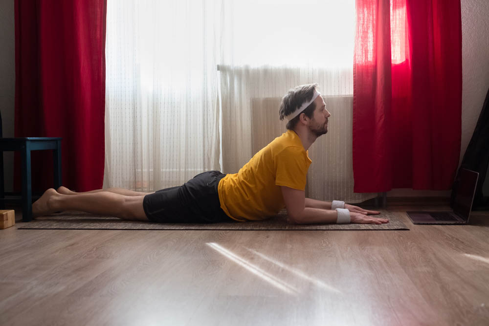 Hombre realizando una postura de yoga acostado boca abajo con la cabeza levantada, vistiendo una camiseta amarilla y muñequeras, en una habitación con cortinas rojas y luz natural entrando.