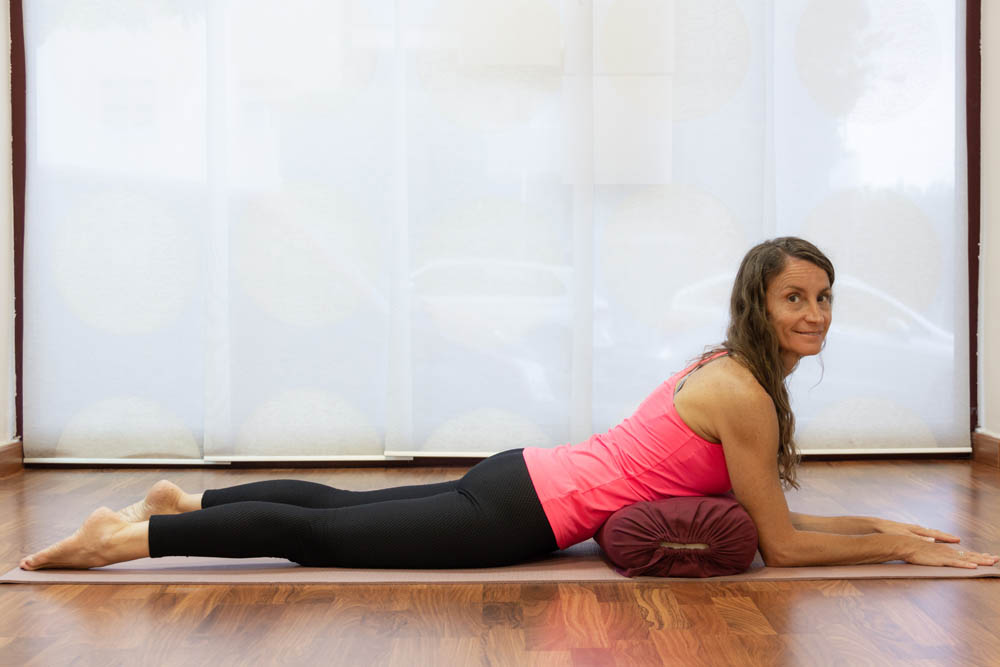 Mujer acostada boca abajo sobre una esterilla de yoga, apoyando los antebrazos en un cojín cilíndrico y mirando hacia la cámara, con luz filtrándose a través de cortinas translúcidas al fondo