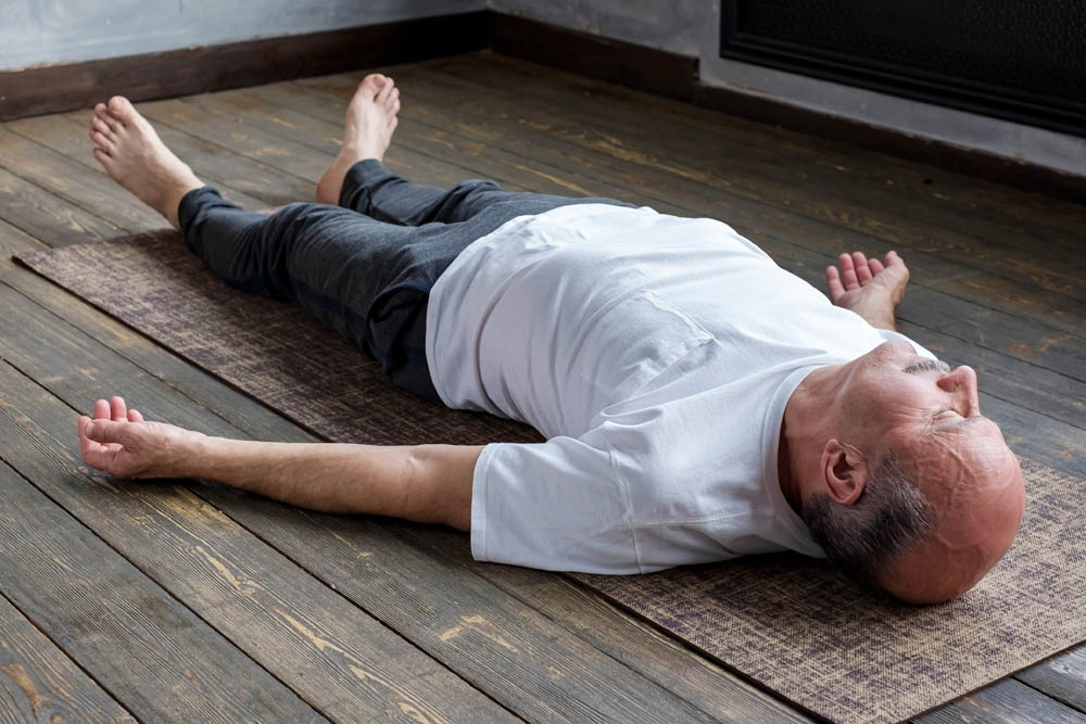 Hombre mayor acostado boca arriba sobre una esterilla de yoga, vestido con camisa blanca y pantalones grises, con los brazos extendidos a los lados y expresión relajada, sobre un suelo de madera