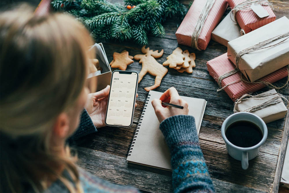 A person writing in a notebook while surrounded by holiday decorations, gift-wrapped presents, and a cup of coffee. The scene conveys a cozy and festive atmosphere.