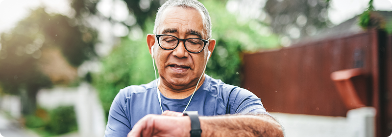 An older man wearing glasses and earphones checks his smartwatch while out for a walk or jog in a suburban neighborhood.