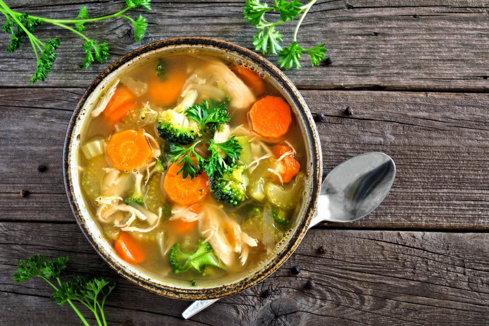 Sopa de verduras con zanahorias, brócoli y apio en un tazón sobre una mesa de madera, decorada con hojas de perejil fresco.