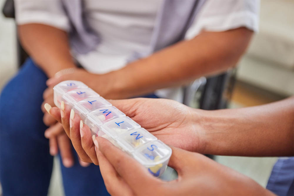 Primer plano de manos sosteniendo un pastillero semanal con compartimentos claramente etiquetados por día. De fondo, otra persona sentada sugiere un entorno enfocado en el cuidado de la salud.