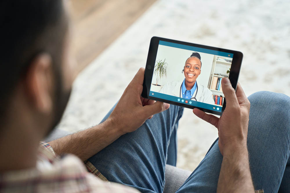 Un hombre sentado con las piernas cruzadas en un sofá sostiene una tablet que muestra una videollamada con un médico