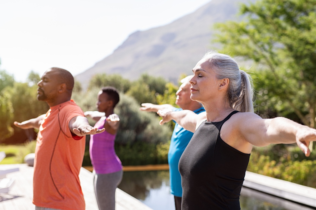 Cuatro personas practicando yoga con los brazos extendidos al aire libre, cerca de una piscina, con árboles y montañas de fondo.