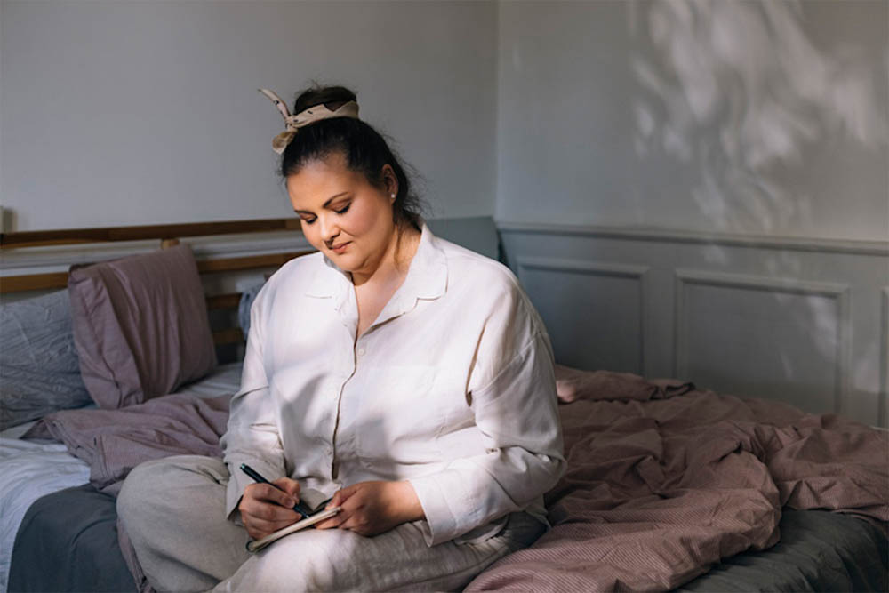 Mujer sentada en su cama con suave luz de la mañana, escribiendo en un diario mientras usa ropa de dormir cómoda.